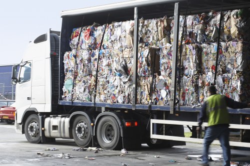 A large flatbed truck parked outdoors, loaded with compressed waste paper and cardboard, which are tightly stacked in large bales. The materials are variously coloured with typical newspaper print, packaging, and brown corrugated cardboard visible. The truck's side panels are open, revealing the bales secured inside. To the right, a worker wearing a high-visibility yellow vest and dark trousers is walking near the truck, with some loose paper on the ground nearby. In the background, part of a parking lot with other vehicles is visible, suggesting an industrial or commercial recycling or rubbish collection site. The scene is lit by natural daylight, emphasizing the textures of the compressed packaging materials and the metallic finish of the truck, illustrating a typical waste removal process for recyclable paper and cardboard in a suburban or urban area, such as Mill Hill in London.