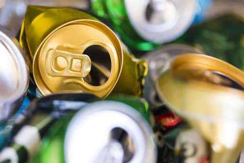 Close-up image of several crushed aluminum cans, including a prominent gold-colored can in the center with an open tab, surrounded by silver, green, and other coloured cans that appear to be flattened and stacked together. The cans are placed on a surface with some greenery visible in the background, suggesting an outdoor setting or a collection area for recyclable rubbish. The metallic surfaces show varying degrees of gloss and texture, with some reflecting light more brightly. The focus is primarily on the open gold can, with the other cans slightly blurred in the background, emphasizing the theme of waste or rubbish collection typical for a professional rubbish removal service such as Garden Clearance Mill Hill, operating around the Mill Hill area, for efficient disposal or recycling of discarded aluminium containers.