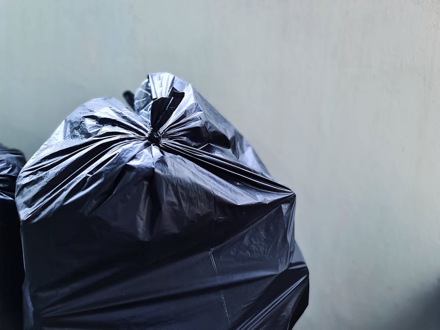 A middle-aged man with short dark hair, dressed in a black t-shirt with white block lettering, is seen engaged in rubbish collection beside a modern, stainless steel public waste bin on a paved sidewa