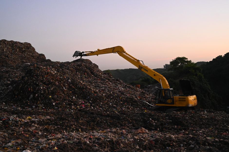 A yellow excavator with a long articulated arm and metal bucket attachment is positioned on a large landfill or waste disposal site during dusk or dawn, with a background of rolling green hills and a 