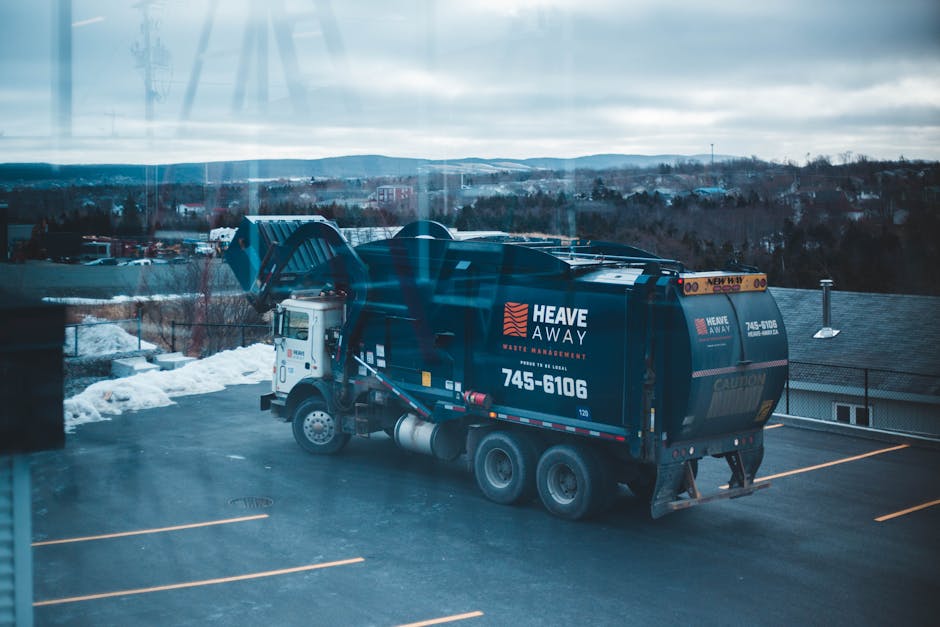 A yellow excavator with a long articulated arm and metal bucket attachment is positioned on a large landfill or waste disposal site during dusk or dawn, with a background of rolling green hills and a 