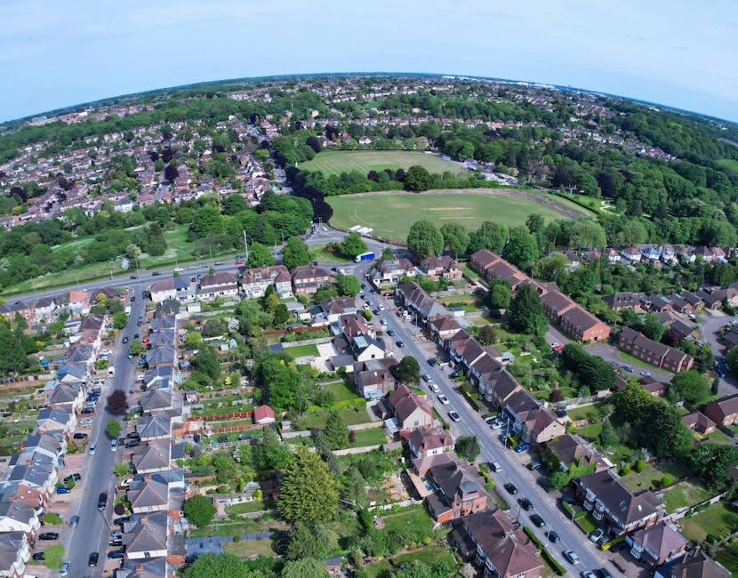 An aerial view of a residential neighbourhood featuring a row of terraced houses with small gardens and driveways in the foreground, constructed with brick and tiled roofs, and a variety of green tree