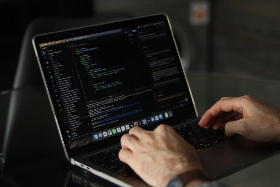A close-up view of a person's hands typing on a laptop with a black keyboard, placed on a dark, smooth surface. The laptop screen displays a dark-themed coding interface with lines of multicolored cod